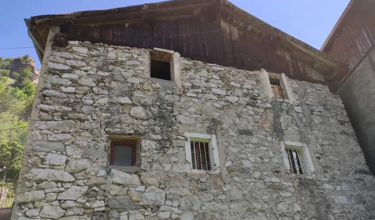 Mur en pierre apparente avec deux fenêtres à volets verts, sous un toit de lauze